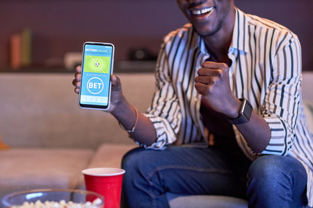 Closeup of black man watching sports match on TV at home and using online betting app