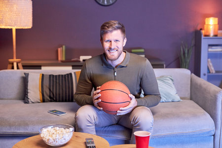 Portrait of emotional man watching sports match on TV at home and holding basketball ball in blue light, copy spaceの写真素材