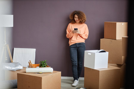 Young woman with curly hair communicating online on smartphone standing among boxes in the room