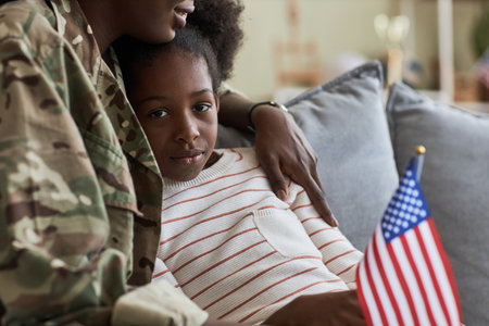 Portrait of African American little girl looking at camera while sitting on sofa together with her mom in military uniformの写真素材