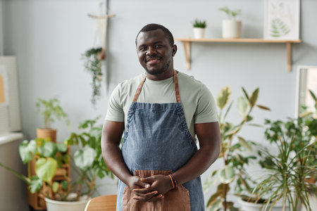 Waist up portrait of smiling black man as gardener wearing apron posing indoors looking at camera with green plantsの写真素材