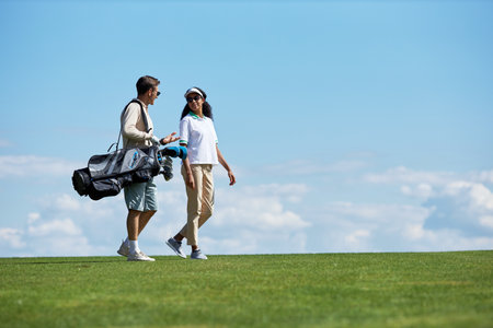 Minimal side view portrait of elegant sporty couple carrying golf bag walking on green field against blue sky, copy spaceの写真素材