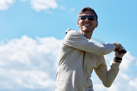 Waist up portrait of smiling man playing golf and swinging golf club against blue sky outdoors, copy spaceの写真素材