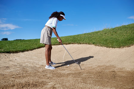 Full length portrait of black young woman playing golf outdoors standing in sandpit and aiming shotの写真素材