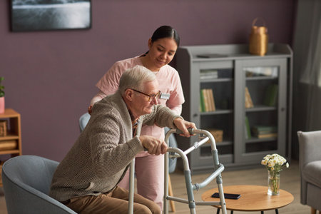 Young social worler caring about man during his rehabilitation, she helping him to stand up with walkerの写真素材