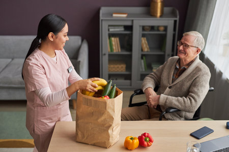Young social worker delivering groceries in paper bag to senior man with disability, she caring about him at homeの写真素材