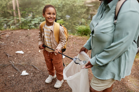 High angle portrait of smiling black girl helping clean nature and picking up plastic bottles outdoorsの写真素材