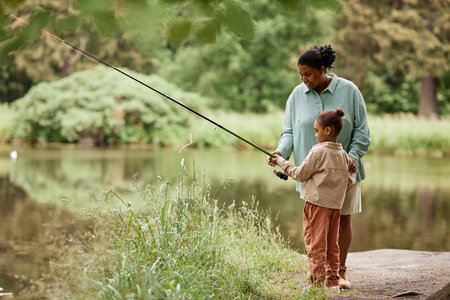 Side view portrait of caring black mother teaching little girl fishing by beautiful forest lake, copy spaceの写真素材