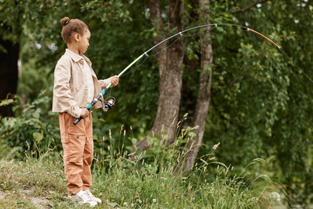 Full length side view portrait of black little girl fishing by river in nature with fishing rod bending, copy spaceの写真素材
