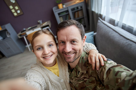 High angle portrait of girl taking photo with father wearing military uniform at home, both looking at camera and smilingの写真素材