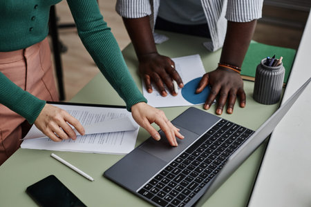 Close up of two people collaborating on project in office and using laptop with documents on table