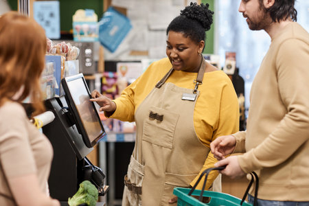 Waist up portrait of smiling black woman helping customers with self checkout in supermarketの写真素材