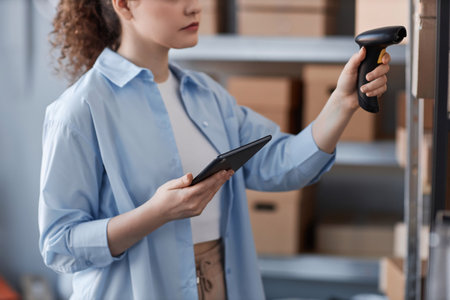 Close-up of young woman in blue shirt scanning qr codes on packed boxes with ordered online goods standing on shelves in storage roomの写真素材