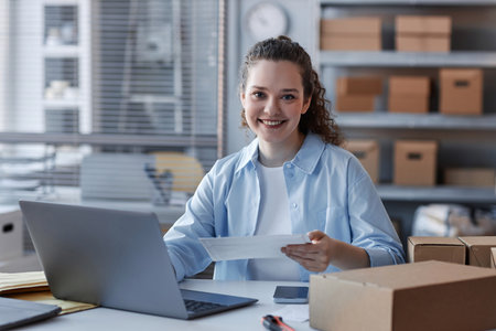 Happy young female manager of marketplace looking at camera while sitting by workplace in front of...