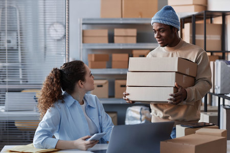 Young black man bringing stack of packed boxes with orders of clients of online shop and looking at female colleague during communicationの写真素材