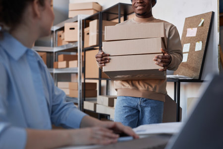 Focus on young black man carrying stack of packed boxes while moving towards his colleague using laptop while taking new online ordersの写真素材