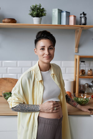 Vertical waist up portrait of tattooed pregnant woman looking at camera while standing in kitchen