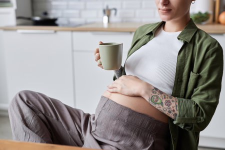 Close up of tattooed pregnant woman relaxing in morning with cup of herbal tea, copy space
