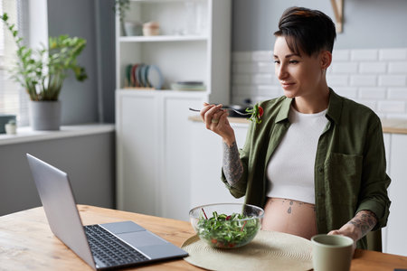 Portrait of modern pregnant woman eating salad in kitchen and watching educational video onlineの写真素材