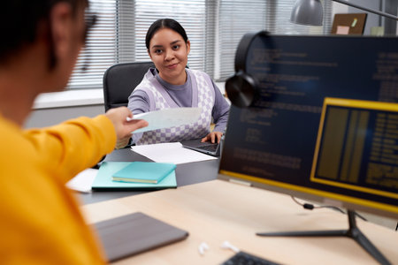Portrait of two young people working in IT development office and handing documents across table...