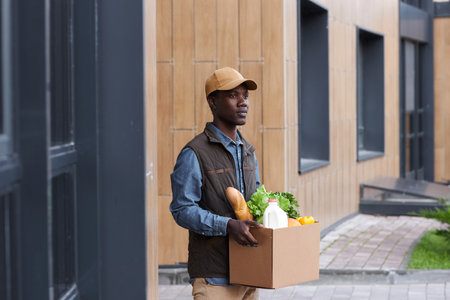 Waist up portrait of black young man carrying box with groceries outdoors while working in food deliveryの写真素材