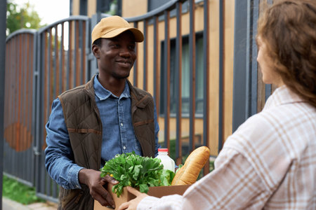 Waist up portrait of black young man handing box with fresh groceries to young woman in cityの写真素材