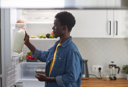 Side view portrait of young black man taking milk from refrigerator while cooking dinner at homeの写真素材