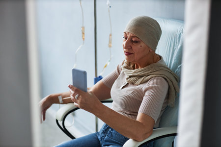 Side view portrait of senior woman sitting in comfortable chair with IV drip and using smartphoneの写真素材
