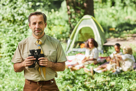 Waist up portrait of mature man as scout leader holding binoculars and looking at camera while...