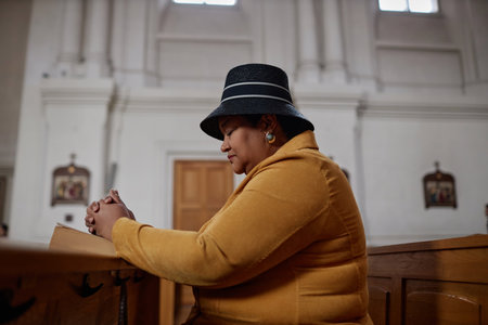 Side view of African American plump woman sitting on bench with Bible and praying in churchの写真素材