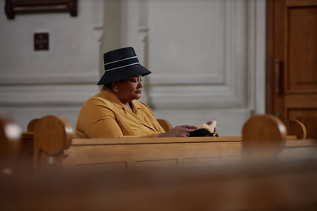 African American woman in elegant clothing reading BIble while sitting on bench in churchの写真素材