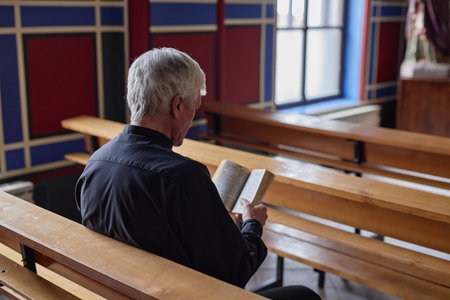 Rear view of senior priest reading Bible during praying while sitting on bench in churchの写真素材