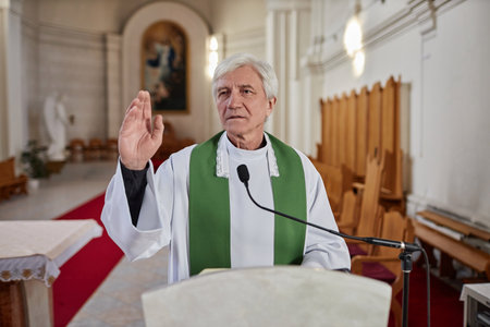 Senior priest in formal wear reading Bible in microphone while standing behind the altar during ceremony in churchの写真素材
