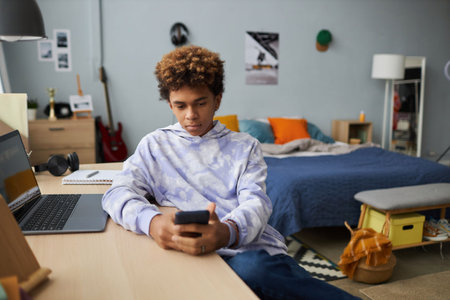 Serious schoolboy in hoodie looking at smartphone screen while watching online video by desk with laptop against double bed with cushionsの写真素材