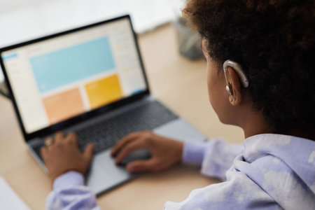 Focus on ear of African American youthful guy in earphones sitting in front of laptop with homepage of website on screen and typingの写真素材