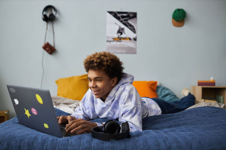 Smiling teenage boy relaxing on bed in front of laptop at leisure and playing games or communicating...