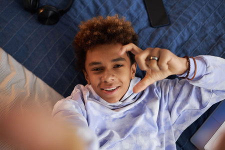 Focus on face of cheerful African American teenage boy looking at camera with smile while relaxing on bed at leisure and taking selfieの写真素材
