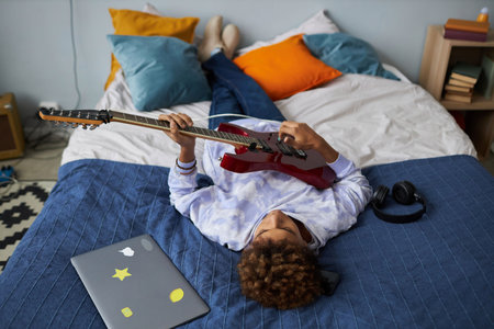Restful African American teenage boy playing electric guitar while relaxing on comfortable double bed after school and enjoying leisureの写真素材