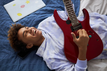 Joyful teenage guy laughing while relaxing on comfortable bed at leisure and playing electric guitar...