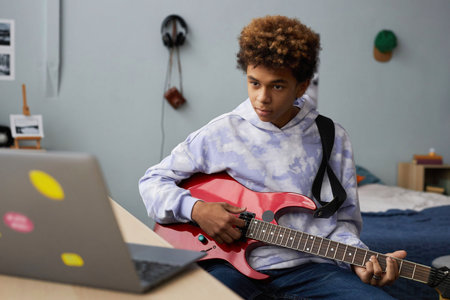 Youthful guy in hoodie sitting on bed at home and looking at laptop screen during online lesson while learning how to play electric guitarの写真素材