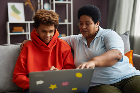 African American mature woman and diligent teenage boy looking at laptop screen while watching...
