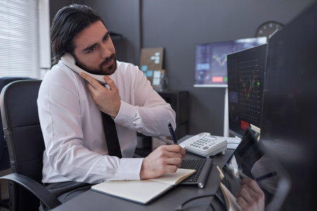 Young broker consulting customer about situation in stock market by the phone sitting at his...