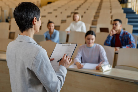 Rear view of teacher reading report to students while they sitting at desk during lecture at universityの写真素材