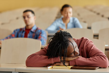 African American schoolboy lying on desk and sleeping during boring lecture with his classmates in...
