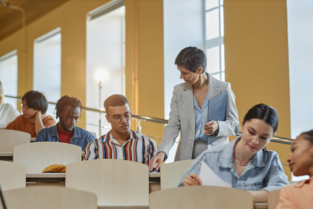 Teacher walking along the lecture hall and giving hints to students during exam while they sitting at desk in a rowの写真素材