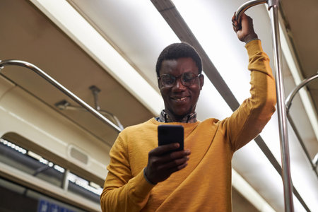 African American man reading message on smartphone and smiling while standing in subway car during journeyの写真素材