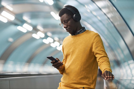 African American man in wireless headphones listening to music on smartphone standing on escalator in subwayの写真素材