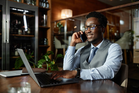 Smiling African American businessman having conversation on mobile phone while working on laptop sitting in cafeの写真素材