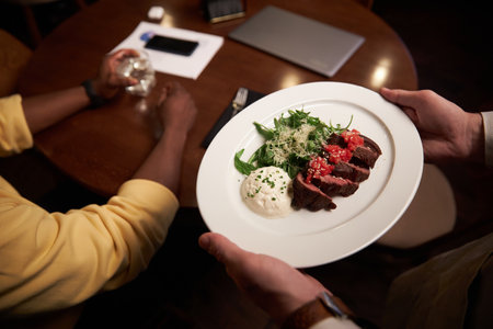 Close-up of waiter carying plate with dinner to customer whil ehe sitting at tableの写真素材