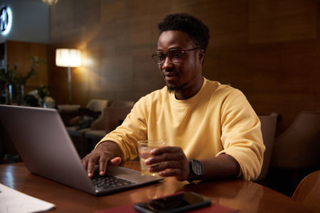 African American man working online on laptop and drinking water while sitting at table in coffee shopの写真素材